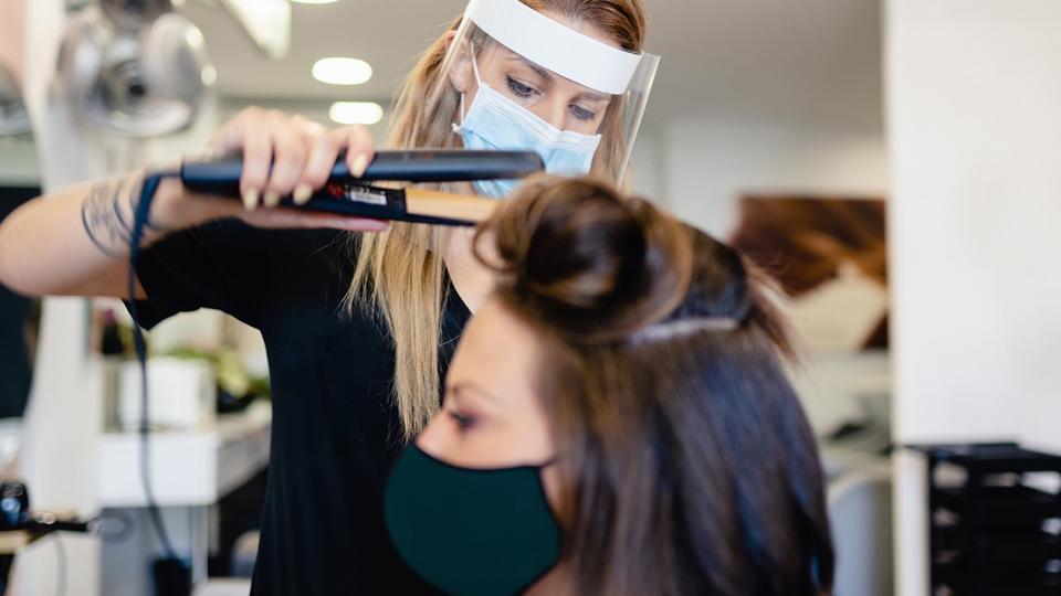 Cosmetology student, wearing a face shield, using a styling tool on client wearing a mask
